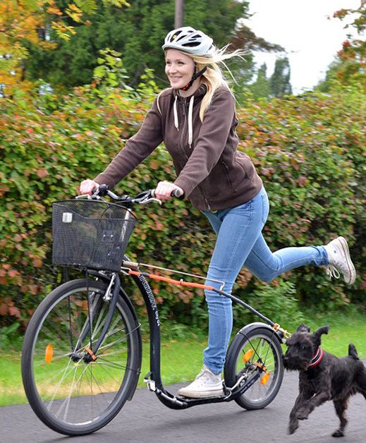 Junge, blonde Frau, die mit einem schwarzen Tretroller fährt. Sie trägt einen weißen Helm und lächelt. neben ihr läuft ein kleiner, schwarzer Hund.