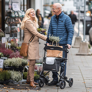 Älterer Mann mit einem Rollator trofft am Blumenladen auf eine Freundin. Beide Lächeln.